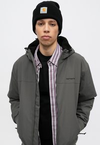 Γκρι τζάκετ με φερμουάρ και μαύρες λεπτομέρειες πάνω από μια μαύρη μπλούζα και ένα πουκάμισο με μοβ ρίγες. Μαύρο πλεκτό σκουφάκι με λογότυπο.