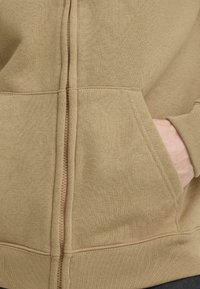 Beige hettejakke med glidelås, glatt tekstur, med sidelommer, ribbestrikkede mansjetter og sømløs glidelåsdetalj på forsiden.