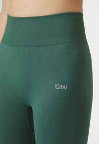 Donker groene high-waisted leggings van glad, rekbaar materiaal. Heeft een geribbelde tailleband en een klein grijs logo op de dij.