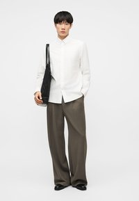 Άνδρας που φοράει λευκό πουκάμισο με κουμπιά, φαρδιά καφέ παντελόνια, μαύρα παπούτσια, κρατώντας μαύρη τσάντα ώμου, στέκεται με το ένα χέρι στην τσέπη.