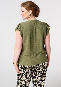 Blouse vert olive avec des manches à volants et un motif texturé, associée à un pantalon noir et crème à imprimé feuillage. Accents de bijoux visibles.