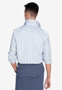Camisa de hombre a rayas con rayas verticales en azul y blanco, de material de algodón, con cuello tipo solapa y mangas enrolladas. Combinada con un suéter de punto azul.