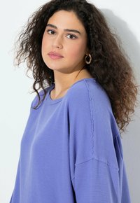 Mujer con cabello rizado oscuro, llevando pendientes de aro dorados y una blusa texturizada de color lavanda, mirando suavemente hacia la cámara contra un fondo blanco.