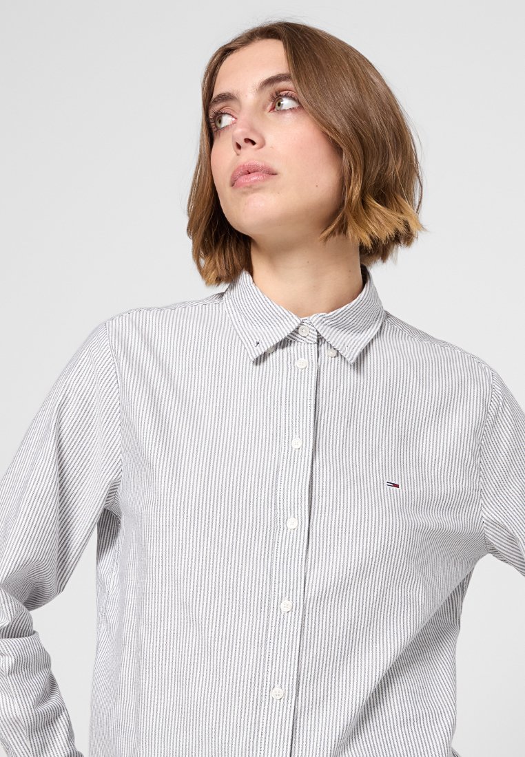 Camicia bianca con bottoni, con un motivo a strisce blu, colletto e chiusura a bottoni. Presenta un piccolo logo sul petto. Il tessuto sembra liscio.