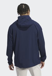 Mężczyzna z krótkimi warkoczykami, ubrany w granatową kurtkę z kapturem i białe spodnie, widziany od tyłu na jednolitym jasnym tle.