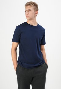 Joven con pelo corto rubio, vistiendo una camiseta azul marino y pantalones gris oscuro, de pie con las manos en los bolsillos, mirando hacia un lado.