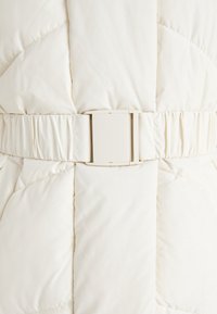 Puffer jacket in cream color with a quilted pattern. Features a wide fabric belt with a rectangular plastic buckle and gathered detailing.