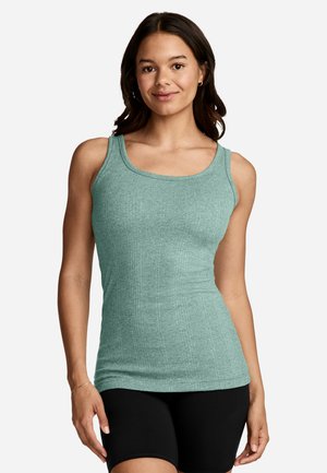 Young woman smiling, wearing a sleeveless light green ribbed tank top and black fitted shorts, standing against a plain background.