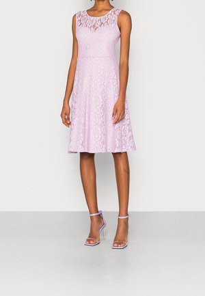 Femme portant une robe en dentelle lilas sans manches avec un décolleté transparent, associée à des sandales lilas à talons hauts, debout devant un fond uni.