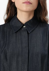 Mørk denimskjorte med spiss krage, én knappelukking og en glatt, lett teksturert overflate. Smal passform med subtil søm.