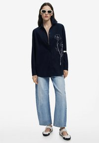Marineblaues Oversize-Hemd mit spitzem Kragen, das auf einer Seite mit weißer Blumenstickerei verziert ist. Kombiniert mit hellblauen, weit geschnittenen Jeans und schwarzen Schuhen.
