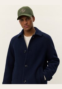 Navy wool coat with a button closure, side pockets, and a collar, paired with a green cap featuring a tonal logo.