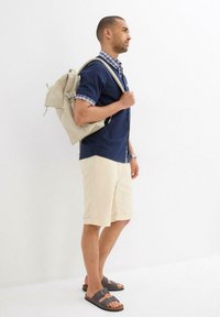 Camicia blu navy a maniche corte con colletto a scacchi, pantaloni corti beige, sandali grigi e uno zaino leggero beige, che presenta un semplice design rettangolare.
