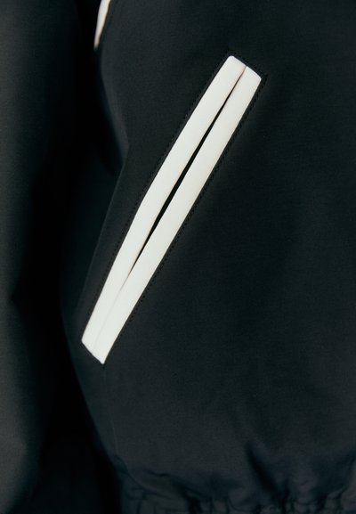 Black jacket with a smooth texture, featuring white accent stripes on the side pockets, structured design, and an elasticated hem.