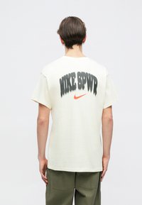 Krēmkrāsas kokvilnas T-krekls ar melnu un oranžu grafisku tekstu mugurpusē, kas lasāms "NIKE SPVR," īsām piedurknēm un apaļu kakla izgriezumu.