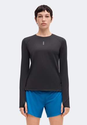 Frau trägt ein schwarzes Langarmshirt mit Daumenlöchern und blaue Sportshorts, steht vor einem einfarbigen hellen Hintergrund.