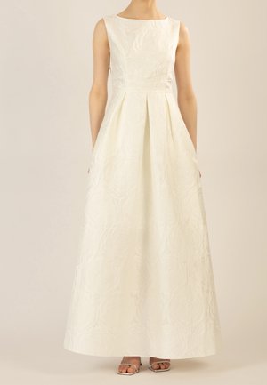 Robe longue texturée sans manches de couleur blanc cassé avec taille cintrée, jupe plissée et encolure carrée, portée avec des talons ouverts argentés.