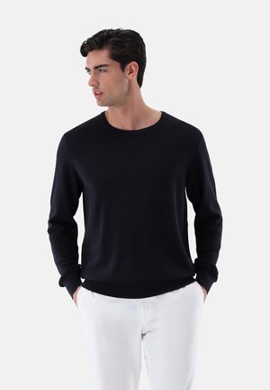 Marineblauer Strickpullover mit rundem Ausschnitt, langen Ärmeln und gerippten Bündchen sowie Saum, getragen über hellen Hosen. Glatte Textur, lässige Passform.