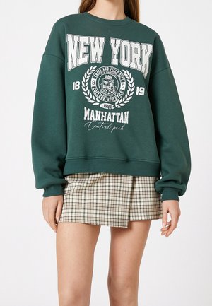Zielona bluza z białym napisem "New York Manhattan Central Park", grafiką emblematu, noszona z beżową kratowaną mini spódniczką na stojącej osobie.