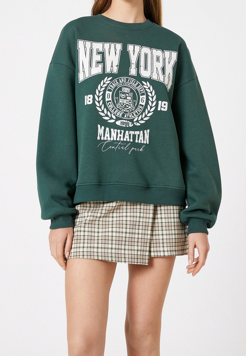Sweat-shirt vert avec texte blanc "New York Manhattan Central Park", motif emblème, porté avec une mini-jupe beige à carreaux sur une personne debout.