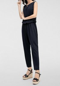 Marinblå overall med ärmlös design, nyckelhålsdetalj vid halsen och elastisk midja. Matchas med svarta kilklackar med remmar.