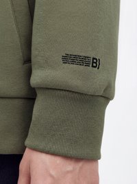 Primer plano del puño de la manga de una sudadera verde con texto impreso en negro y un diseño similar a código cerca de la muñeca, sobre una mano visible.