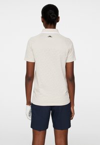 Kortärmad beige golfpolo med texturerad baksida, vit krage och svart logga på nacken. Kombinerad med marinblå shorts.