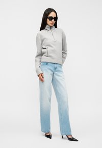 Γκρι πουλόβερ με φερμουάρ στον λαιμό και πλαϊνή τσέπη, σε συνδυασμό με ανοιχτό μπλε τζιν παντελόνι με φαρδιά μπατζάκια και μαύρα τακούνια με μυτερή μύτη. Ολόκληρο το ντύσιμο έχει ομαλές υφές.