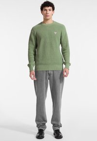 Maglione verde a coste con scollatura rotonda e piccolo logo, abbinato a jeans grigi a gamba dritta e scarpe nere.