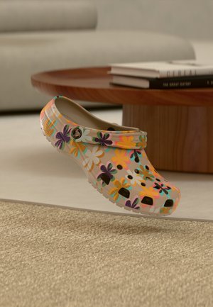 Beżowy chodak z jasnym, wielokolorowym kwiatowym wzorem, otworami wentylacyjnymi i paskiem, unoszący się nad beżowym dywanem w salonie.