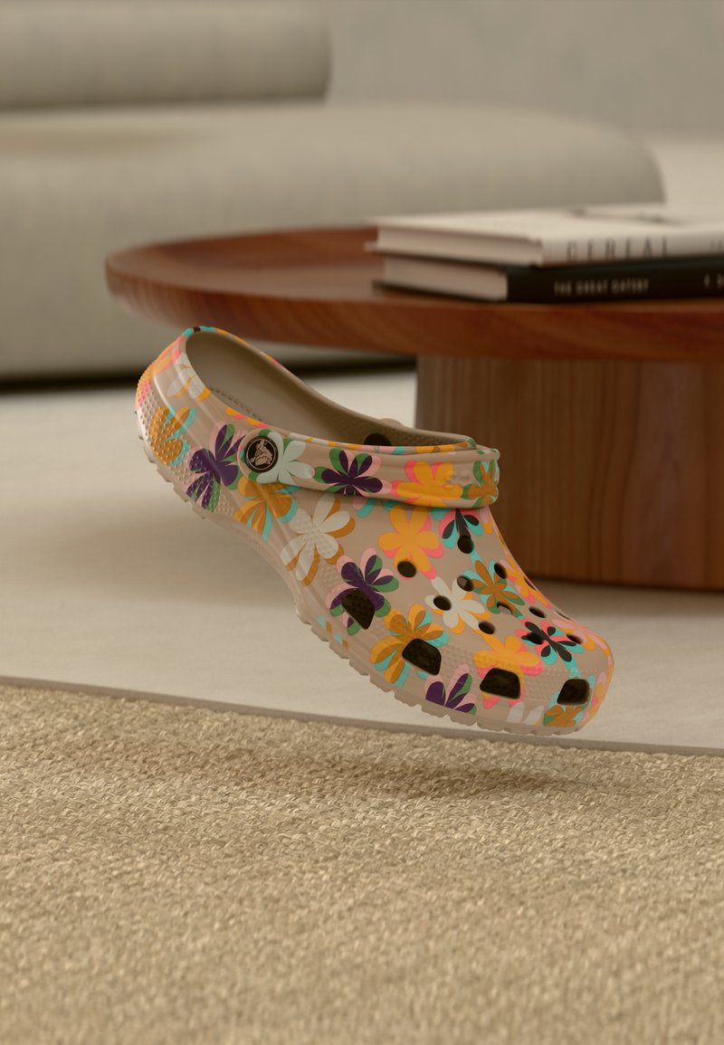 Μπεζ κλογκ με φωτεινό πολύχρωμο λουλουδάτο μοτίβο, οπές αερισμού και ιμάντα, να αιωρείται πάνω από ένα μπεζ χαλί σε σαλόνι.