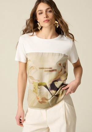 Kvinde med langt brunt hår iført hvid top med beige blomsterprint og teksten "exist in moments, nothing more," kombineret med hvide bukser.