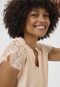 Blusa rosa pallido con maniche corte in pizzo, scollatura arricciata e un sottile dettaglio a chiave sul retro. Tessuto liscio con una delicata texture.