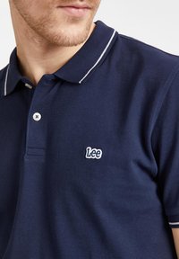 Granatowa koszulka polo z teksturowanej tkaniny, z dwoma guzikami przy dekolcie, białymi zdobieniami kołnierzyka oraz małym białym haftowanym logo "Lee".