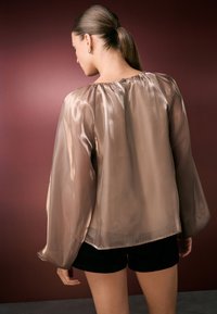 Kerge, helepruun pluus pika puhvärvega ja kogutud kaelusega, millel on sile tekstuur ja tagumine nööpdetail, koos mustade lühikeste pükstega.