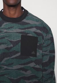 Personne portant un t-shirt à manches longues avec un motif camouflage vert et gris, avec une poche poitrine noire avec un logo Adidas.
