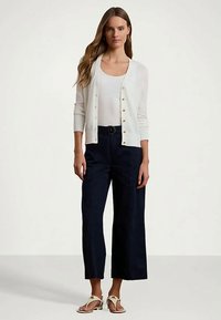 Sieviete stāv, ģērbusies baltajā pogātajā kardiganā virs baltas blūzes, melnās jostu plašajās biksēs un baltos sandalos uz vienkārša fona.