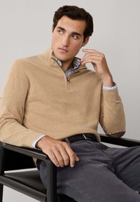Giovane seduto su una sedia nera, indossa un maglione beige con zip fino al quarto sopra una camicia a righe blu e pantaloni grigi, mentre guarda pensieroso di lato.