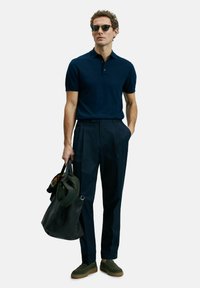 Polo-shirt marine, pantalon marine ajusté, chaussures vertes à enfiler, portant un sac noir avec des accents verts. Textures lisses, coupe décontractée.