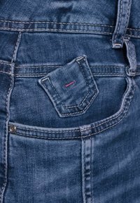 Blå denim jeans med en liten, rektangulär ficka som har röd stygning. Slät yta med synlig sömnad och blekta områden.