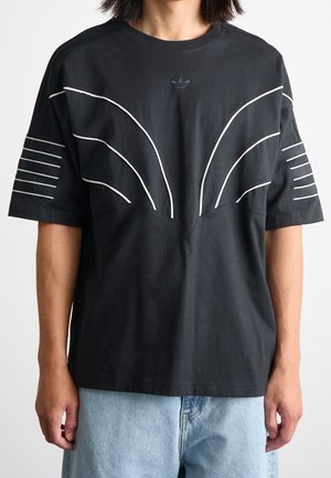 Czarna, oversize'owa koszulka z białymi, zakrzywionymi i paskowanymi liniami, logo Adidas na piersi, noszona z jasnoniebieskimi dżinsami.