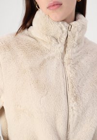 Faux fur jacket in cream with a high collar and zip closure. Soft texture with a smooth lining and subtle sheen.