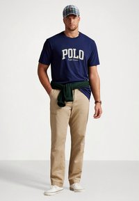 Μπλε ναυτικό βαμβακερό t-shirt με το κείμενο "POLO", μπεζ βαμβακερό παντελόνι και πράσινο πουλόβερ δεμένο στη μέση. Καρό καπέλο και λευκά αθλητικά παπούτσια.