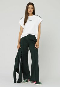 T-shirt blanche à manches courtes avec du texte noir, associée à un pantalon vert foncé. Le modèle tient une veste verte assortie et porte des baskets vertes avec des accents roses.