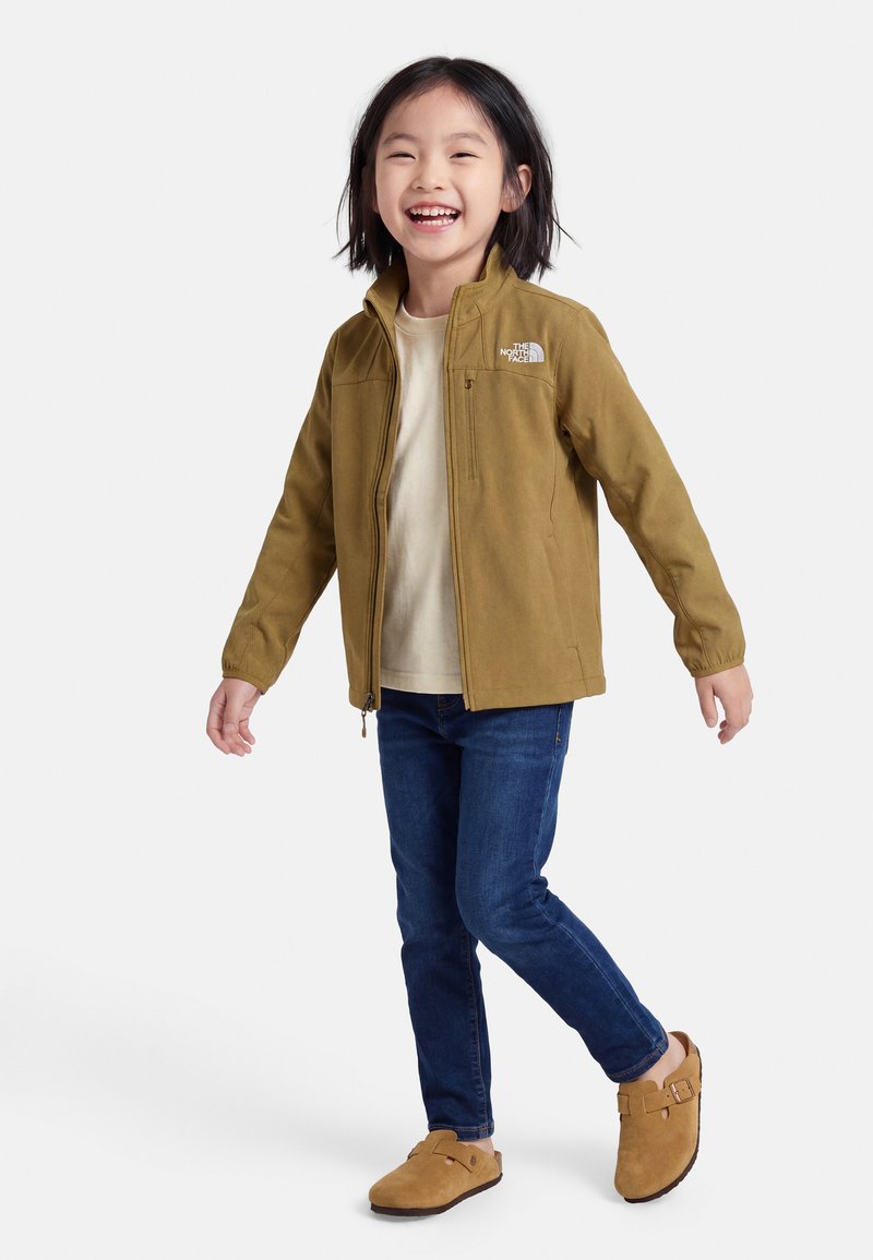 Χαμογελαστό παιδί που φοράει καφέ ζακέτα με φερμουάρ, μπεζ πουκάμισο, μπλε τζιν και καφέ παπούτσια χωρίς κορδόνια, όρθιο με το ένα πόδι ελαφρώς σηκωμένο.