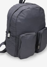 Mochila gris con un acabado suave y texturizado. Cuenta con dos bolsillos frontales con cremallera y correas de hombro acolchadas. Silueta redondeada.