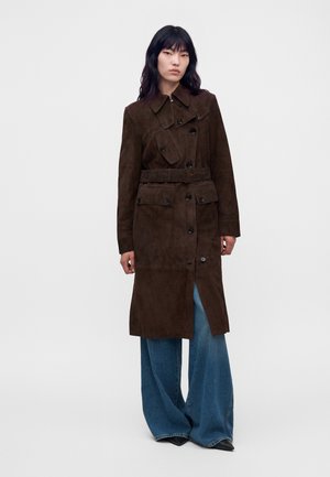 Une femme porte un long manteau marron foncé ceinturé avec des poches à rabat, par-dessus un jean bleu à jambes larges, sur un fond d’étude uni.