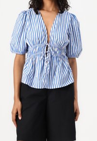 Blau-weiß gestreifte Bluse mit tiefem V-Ausschnitt, Puffärmeln und geraffter Taille mit Kordelzugdetail. Kombiniert mit schwarzen Shorts.