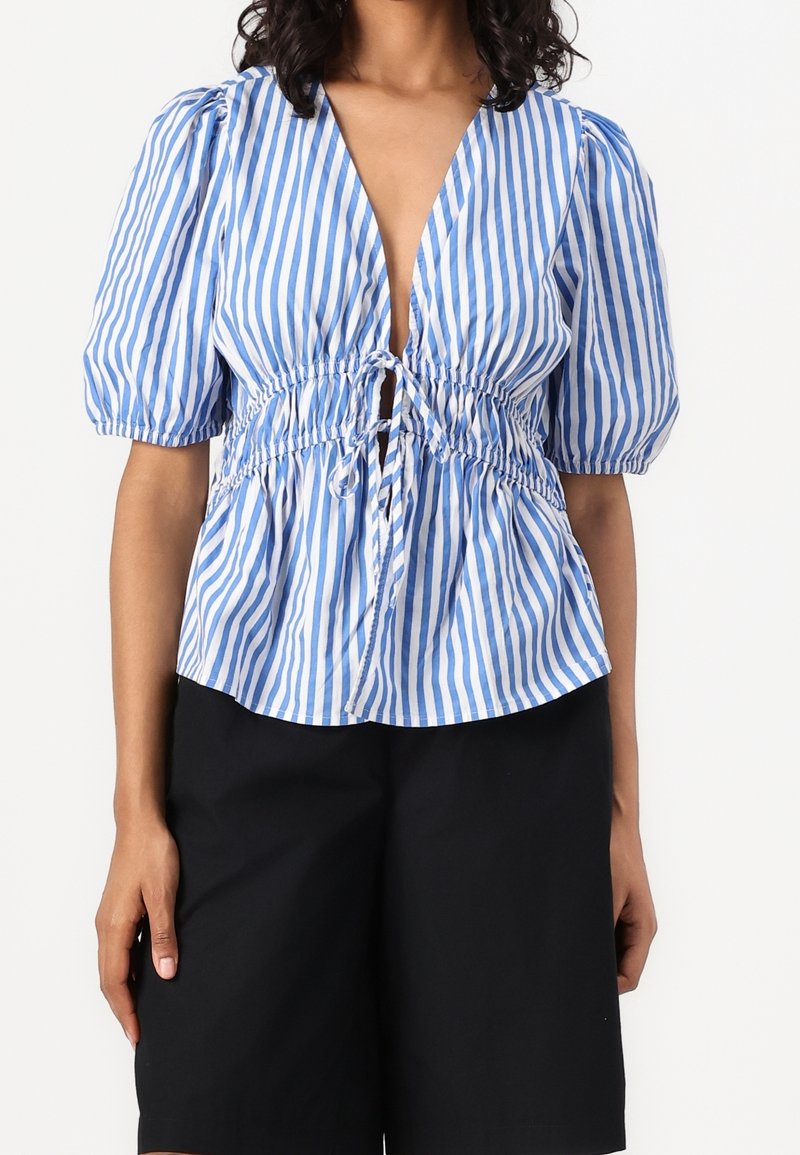 Blau-weiß gestreifte Bluse mit tiefem V-Ausschnitt, Puffärmeln und geraffter Taille mit Kordelzugdetail. Kombiniert mit schwarzen Shorts.