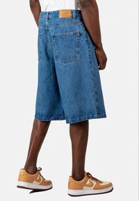 Chándal de mezclilla en azul claro con un corte relajado, que cuenta con dos bolsillos traseros y un parche de cuero marrón en la cintura. Combinado con zapatillas beige.